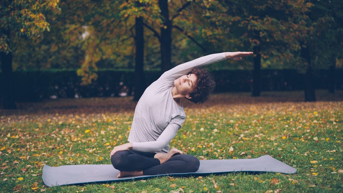 Slim young lady is doing sequence of yoga asanas sitting on mat in park alone on warm autumn day with beautiful grass and trees around her. Recreation and sport concept.