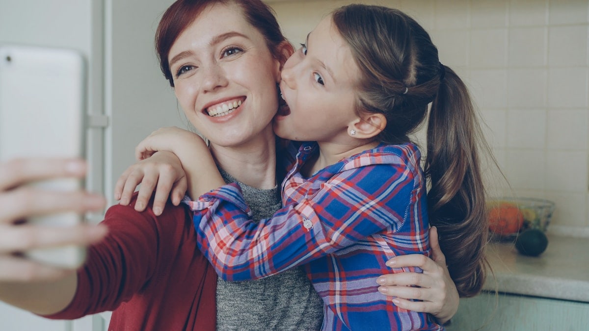 Smiling mother together with funny playful daughter making selfie photo with smartphone camera at home in kitchen. Family, cooking, and people concept